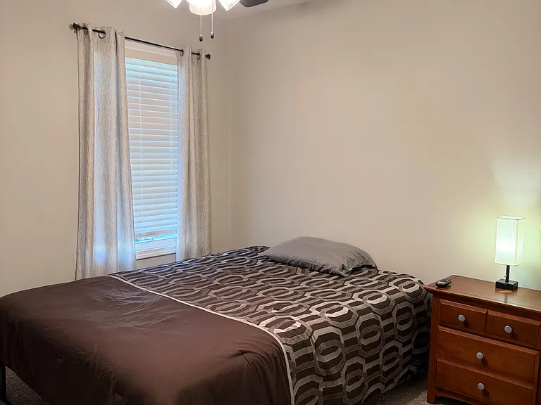 Comfortable queen bedroom featuring a ceiling fan with light, a window with blinds and drapes, and a nightstand with lamp.
