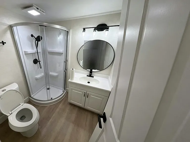 Clean bathroom with corner shower enclosure, vanity with storage, and round mirror under updated lighting.