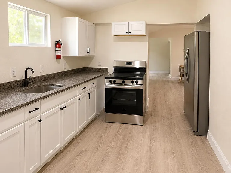 Galley kitchen with ample white cabinetry, stone-look counters, stainless steel appliances, and a window over the sink.