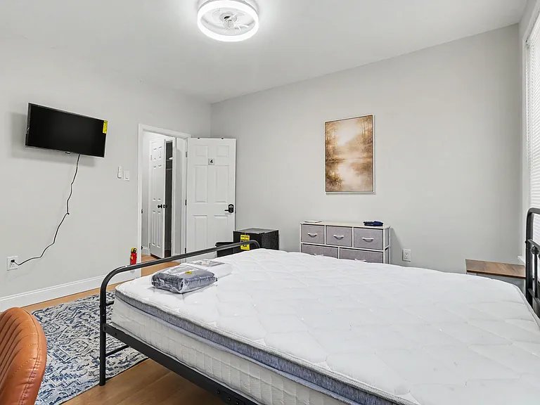 Furnished bedroom with a full bed, dresser storage, and wall-mounted TV, plus a window with blinds for natural light.