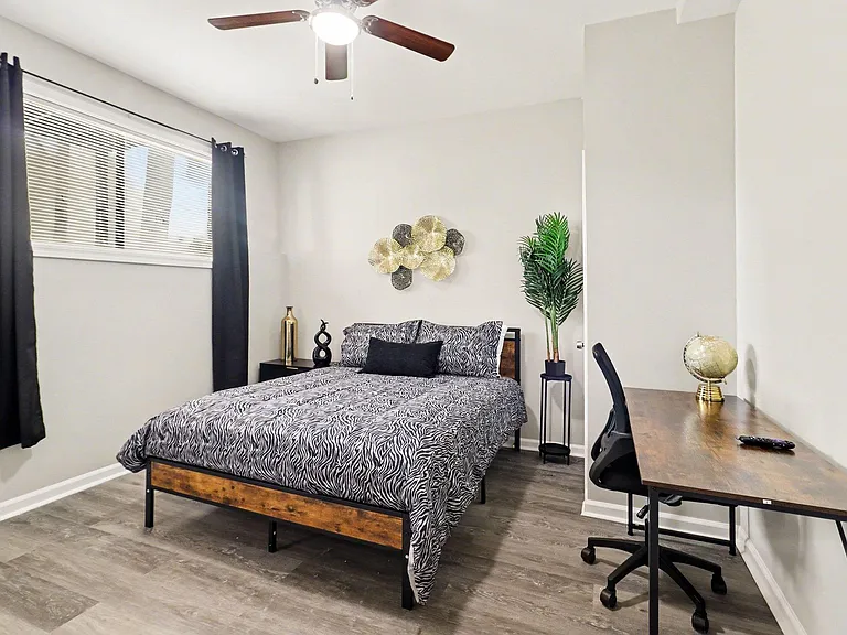 Private bedroom with a queen bed, large window with curtains, ceiling fan, and dedicated desk workspace on wood-look floors.