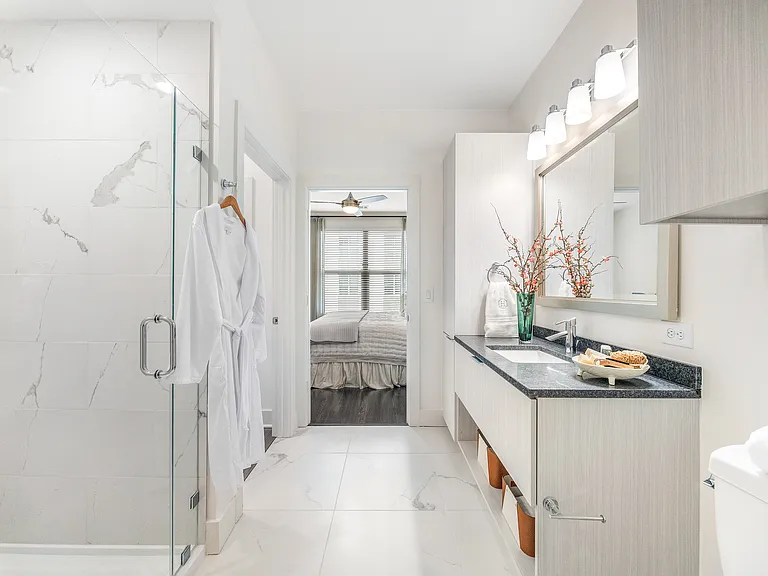 Elegant bathroom featuring a luxurious shower, plush white robe, and modern vanity with stylish decor, leading to a cozy bedroom with natural light.