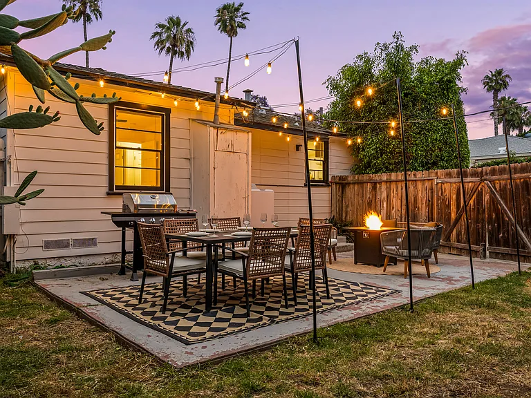 Cozy backyard retreat with cafe-style string lights, outdoor dining, a BBQ grill, and a fire pit lounge. Surrounded by palm trees and warm lighting, this inviting space is perfect for relaxing evenings and group gatherings.