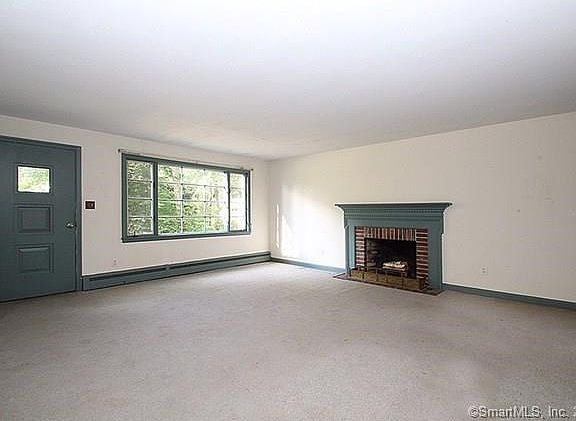 Large living room with fireplace and large front window. There is hardwood flooring under the carpet.
