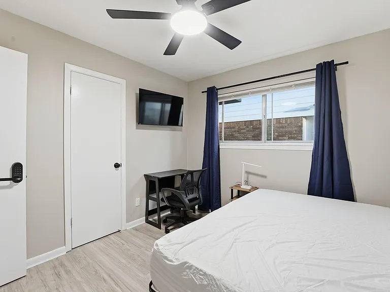 Private bedroom with a ceiling fan, wall-mounted TV, work desk, and a large window with curtain panels for light control.
