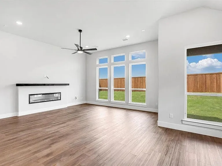 Spacious living area featuring wood-finish flooring, a linear electric fireplace with a dark mantel, and multiple large windows overlooking the backyard