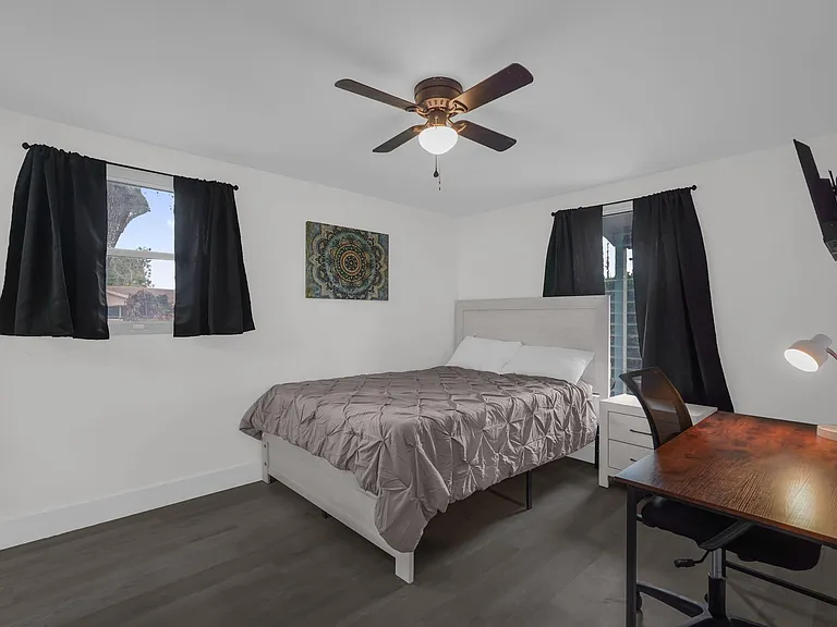 Furnished bedroom with a full bed, nightstand, and dedicated desk area, plus ceiling fan and window coverings.