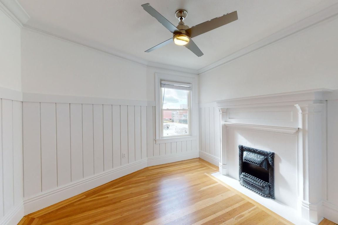 Bright room with hardwood floors, paneled walls, a window, and a decorative fireplace, modern ceiling fan above