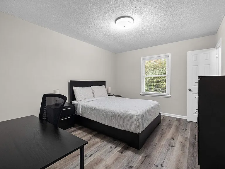 Furnished bedroom with a full bed, nightstands, and a dedicated desk area, plus a window and closet for storage.