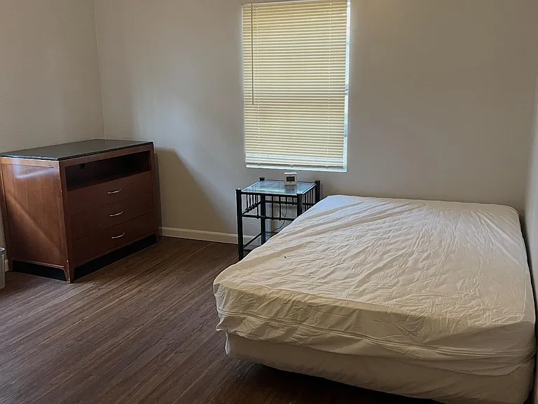 Furnished bedroom with wood-look flooring, a twin bed, dresser, and nightstand, plus a window with blinds for privacy.