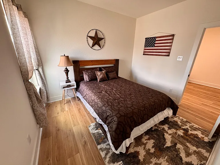Furnished bedroom with a queen bed, wood-style flooring, window with curtains, and a nightstand with lamp.