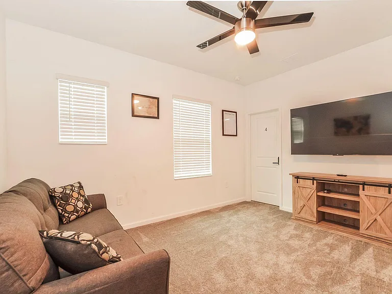 Carpeted living area with ceiling fan, dual windows with blinds, and a wall-mounted TV with media console for easy lounging.