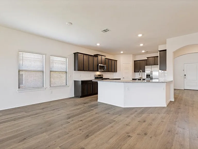 Open-concept living space with wood-finish flooring, a kitchen island, dark wood cabinetry, stainless steel appliances, and two windows with blinds