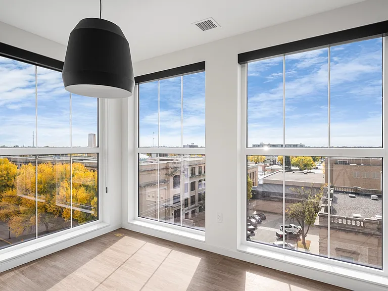 Bright corner room with large windows offering stunning urban and tree-lined views, complemented by a modern hanging lamp. Bright corner room with large windows offering stunning urban and tree-lined views, complemented by a modern hanging lamp.