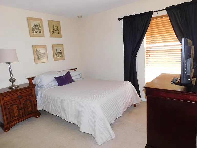 Furnished bedroom featuring a comfortable bed, nightstand with lamp, dresser with TV, and window blinds for light control.