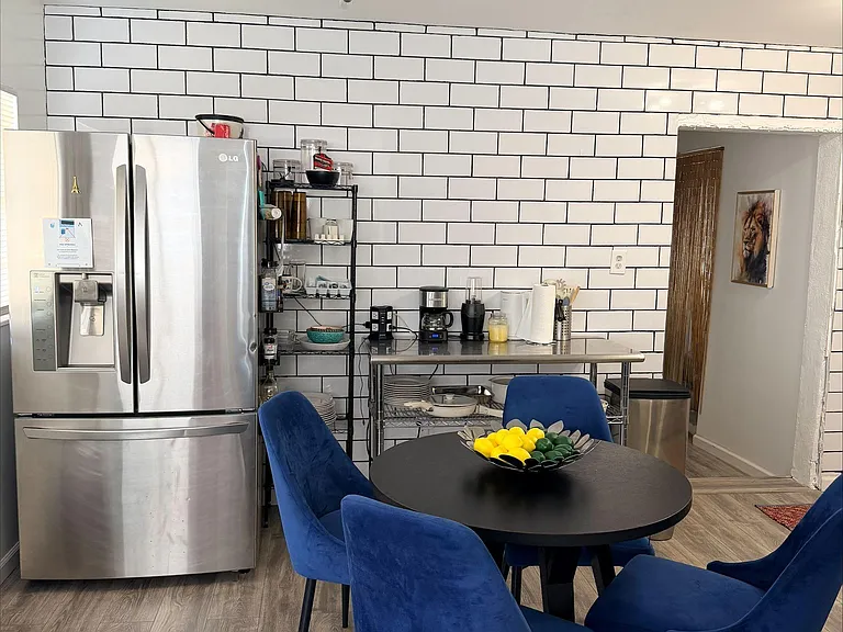 Eat-in kitchen area with round dining table, stainless-steel fridge, open shelving, and easy-clean tile-style accent wall.