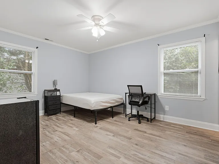 Spacious furnished bedroom with two windows, ceiling fan, and wood-look flooring plus a desk and dresser for storage.