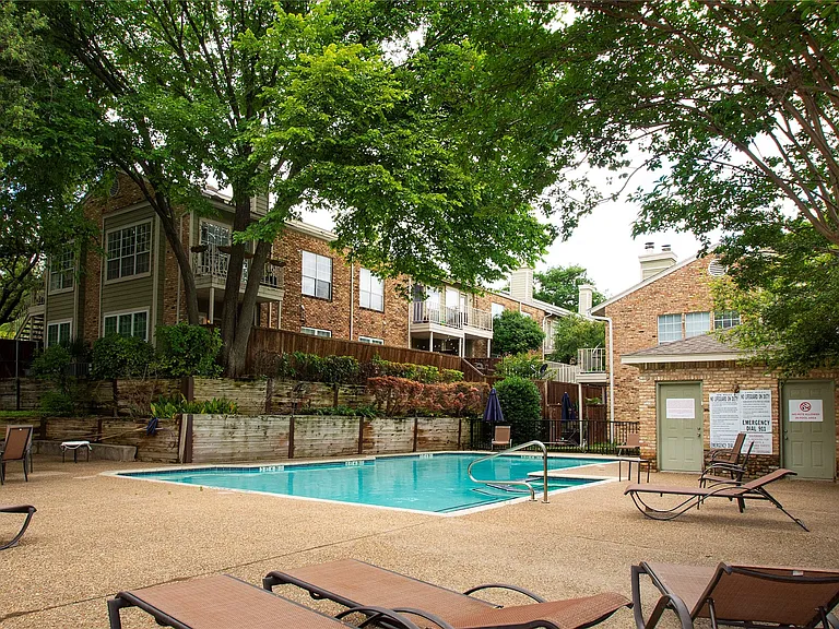 Community swimming pool with a textured concrete deck, surrounded by mature trees, brick-clad buildings, and wooden retaining walls