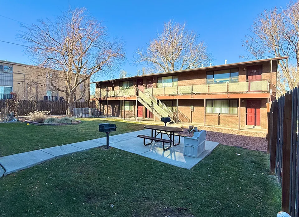 Cozy apartment complex with charming red doors and an inviting courtyard featuring a picnic area and barbecue spot, surrounded by lush greenery.