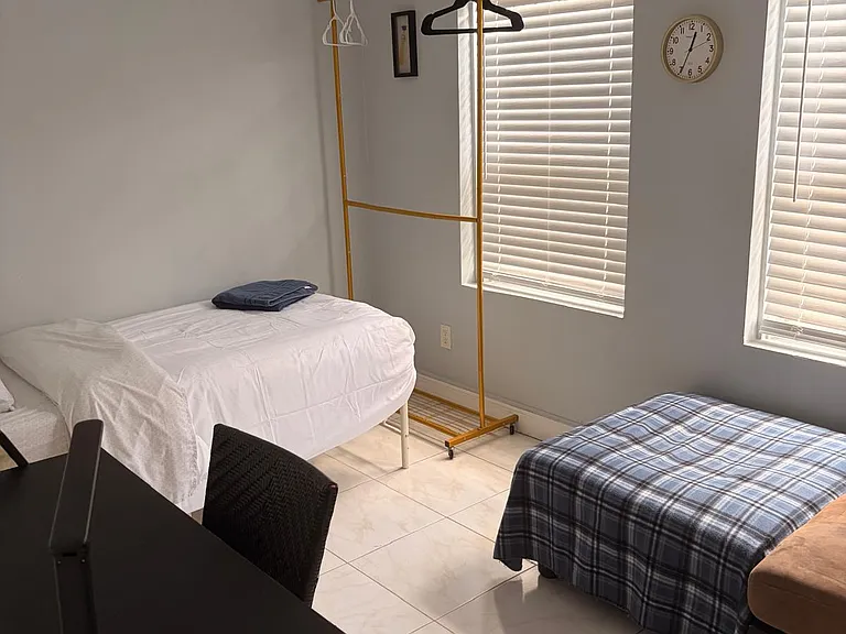 Private bedroom with tile flooring, two windows with blinds, and space for a bed plus a small work area and wardrobe rack.