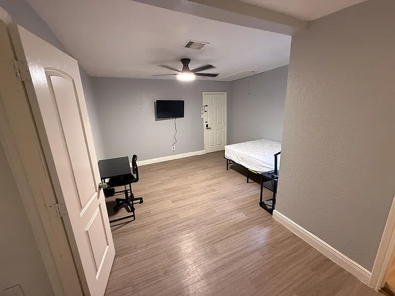 Furnished bedroom with wood-look flooring, ceiling fan, wall-mounted TV, and a work desk for studying or remote work.