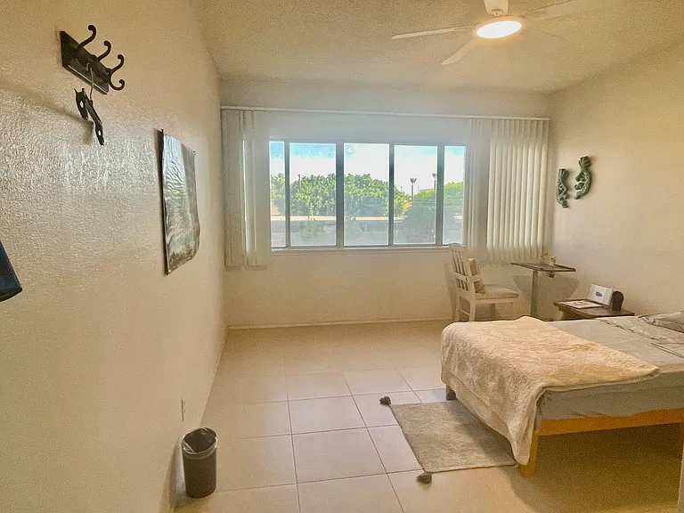 Bedroom with a large window and vertical blinds, tile flooring, and space for a bed plus a small desk or seating area.
