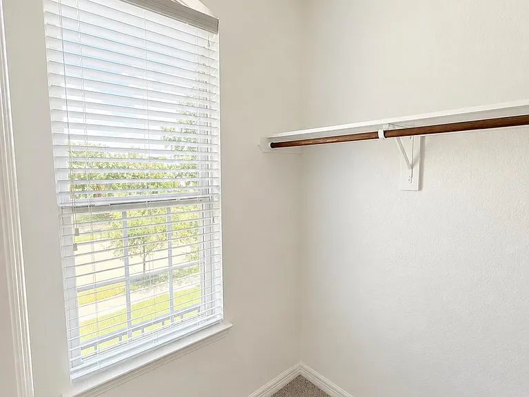 Carpeted walk-in closet with a hanging rod and shelf, plus a window with blinds for natural light and ventilation.