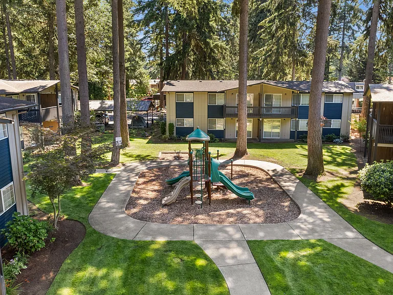 A beautifully landscaped apartment courtyard featuring a playground, mature trees, walking paths, and peaceful outdoor green space.