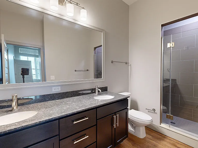 Apartment bathroom with glass-enclosed shower, wood-style flooring and double sinks.