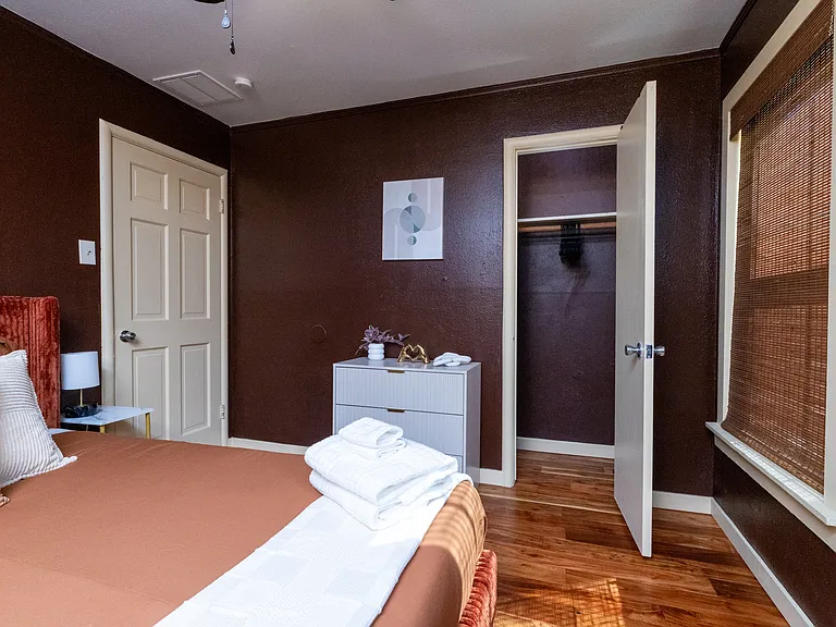 Cozy retreat with warm tones and natural light. This comfortable bedroom features a queen bed, bamboo shades, a ceiling fan, and a modern mirror accent _ perfect for unwinding after a day exploring Dallas.