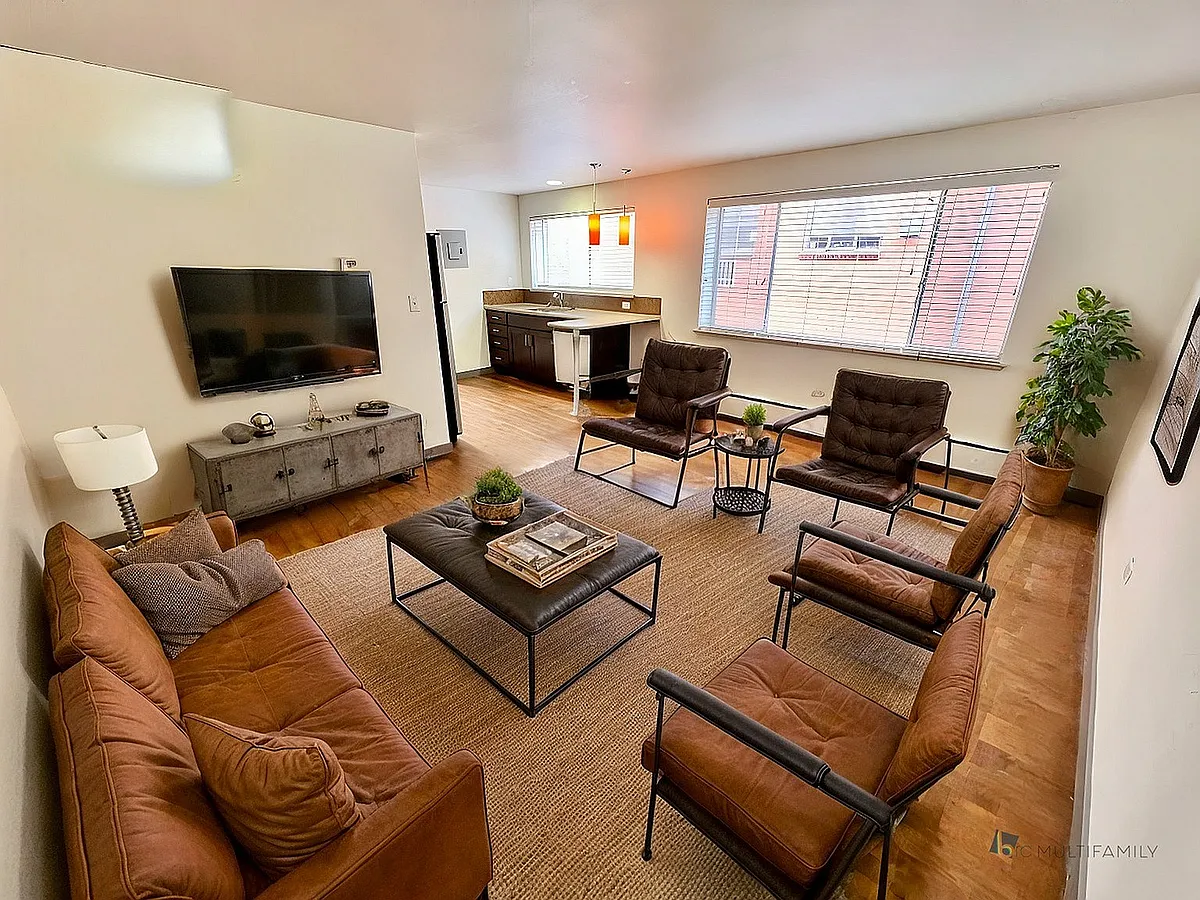 Modern living room with stylish brown leather seating, a mounted flat-screen TV, natural light from large windows, and open access to a sleek kitchen. - Patina