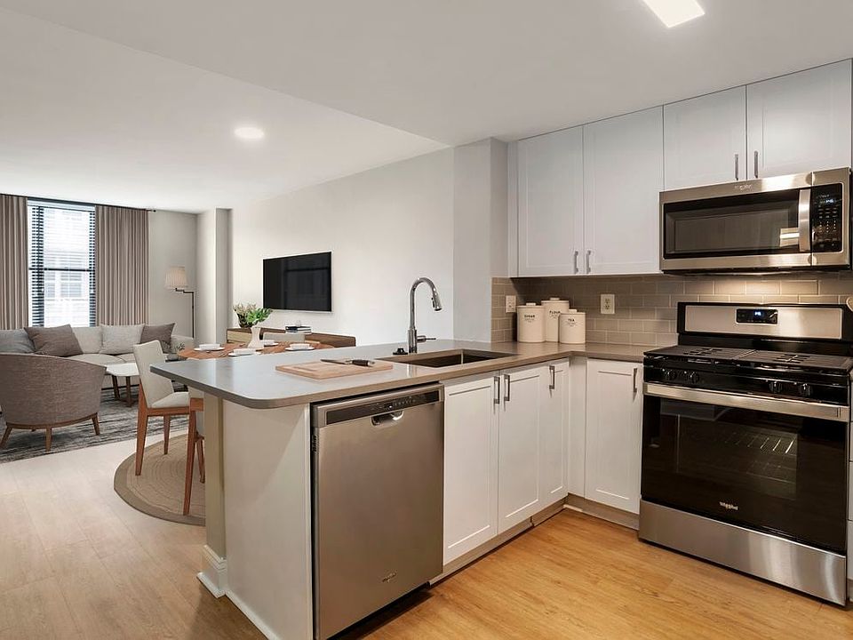 Renovated Package I kitchen and living area with white cabinetry, grey quartz countertops, stainless steel appliances, grey tile backsplash, and hard surface flooring