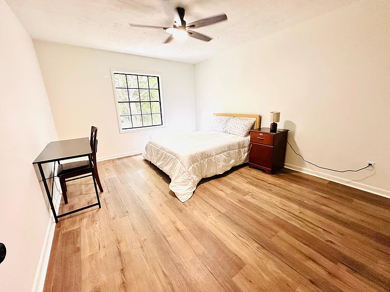 Spacious furnished bedroom with wood-style flooring, ceiling fan, and a large window, plus a desk and nightstand.