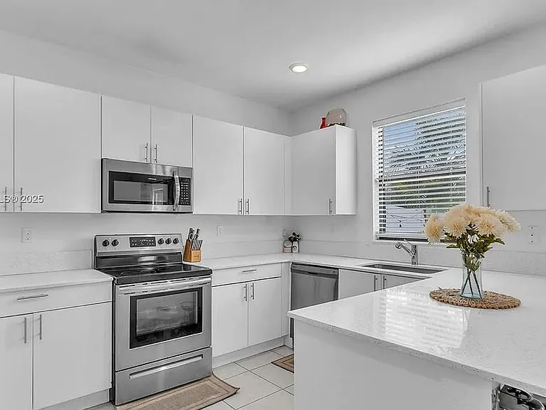 Modern all-white kitchen with quartz counters, stainless steel appliances, and ample natural light through a window above the