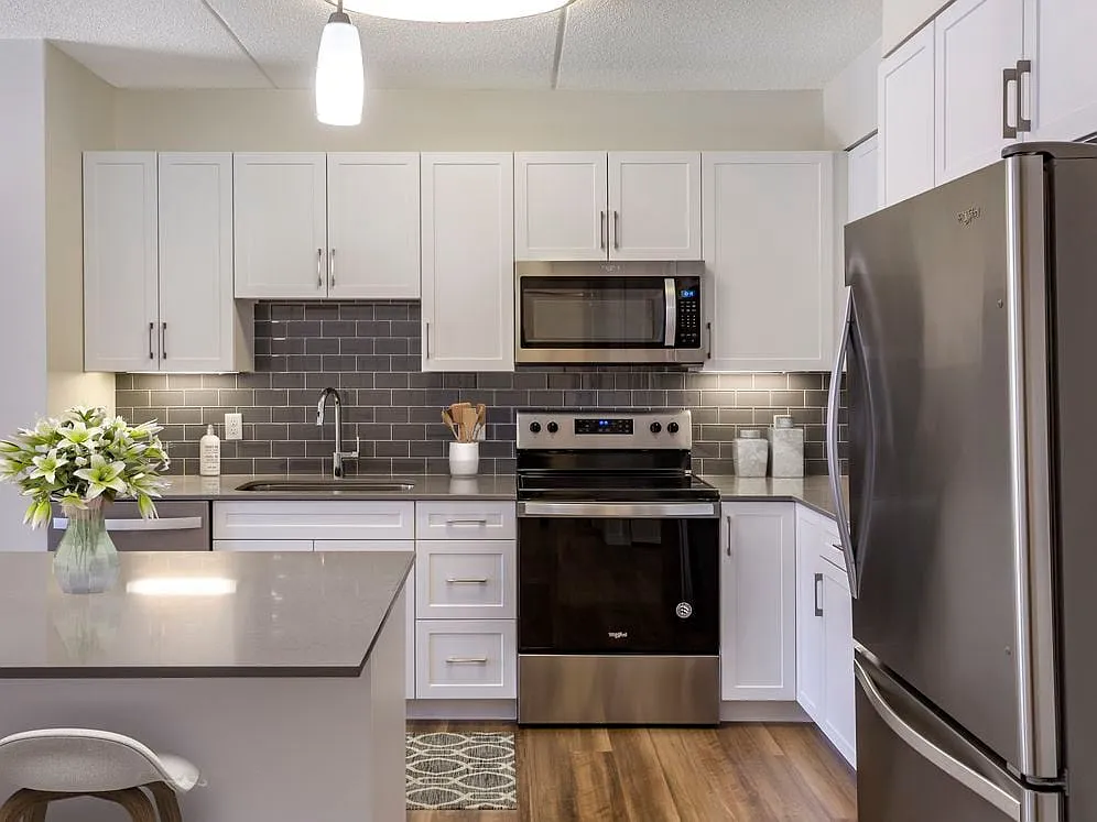 Renovated Package II kitchen with grey quartz countertops, white shaker cabinetry, stainless steel appliances, grey tile backsplash, and hard surface flooring throughout - Avalon Natick