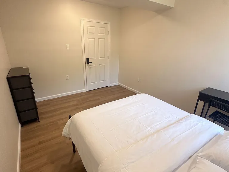 Private furnished bedroom with bed, desk setup, and shelving, featuring durable wood-look flooring and a secure entry door.