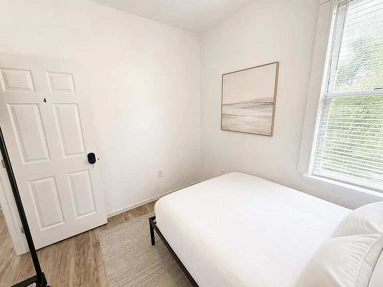 Private bedroom with a full-size bed, wood-look flooring, and a large window with blinds for natural light and privacy.