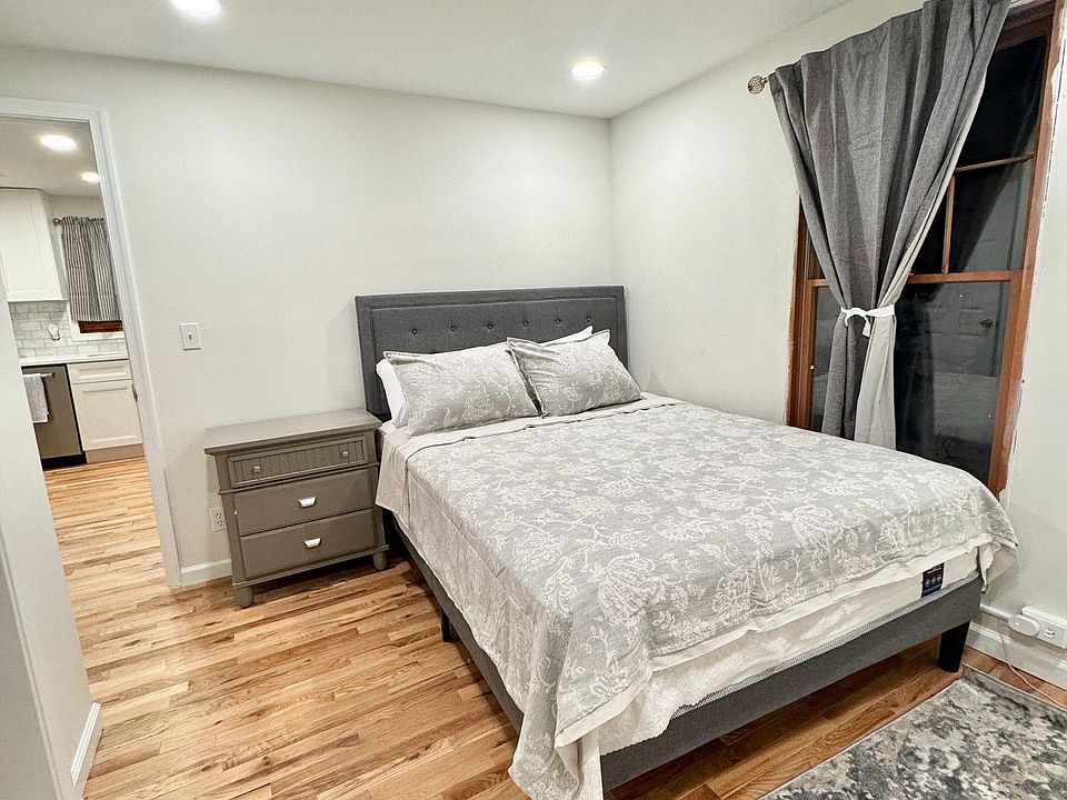 Furnished bedroom with a full bed, nightstand, hardwood-style floors, and a window with curtains for privacy.