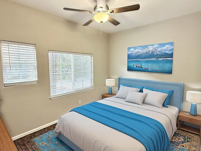 Inviting bedroom with a large bed featuring blue accents, modern decor, natural light from two windows, and a serene mountain landscape art piece.