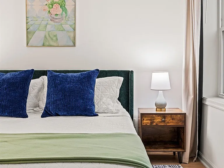 Furnished bedroom with a comfortable bed, nightstand with lamp, and window with curtains for natural light.