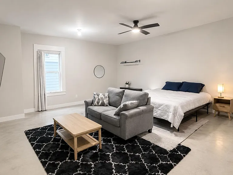 Spacious furnished bedroom with a king bed, seating area, wall-mounted TV, and ceiling fan, plus a window with curtains.