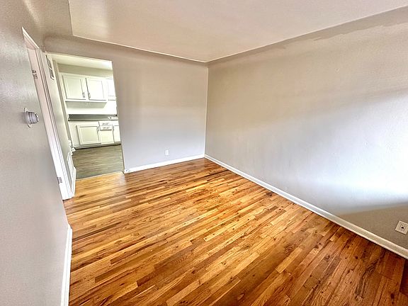 Bright, open room with hardwood floors leading to a modern kitchen with sleek white cabinetry.