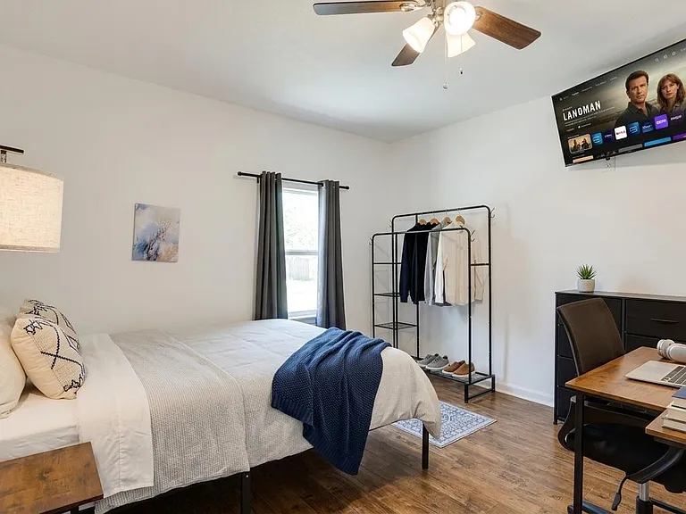 Spacious bedroom with a full bed, work desk, wall-mounted TV, ceiling fan, and window with curtains for privacy.