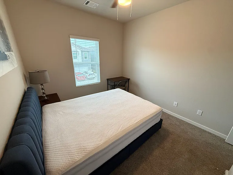 Private furnished bedroom with a full-size bed, nightstand and lamp, plus a compact desk by the window for study.