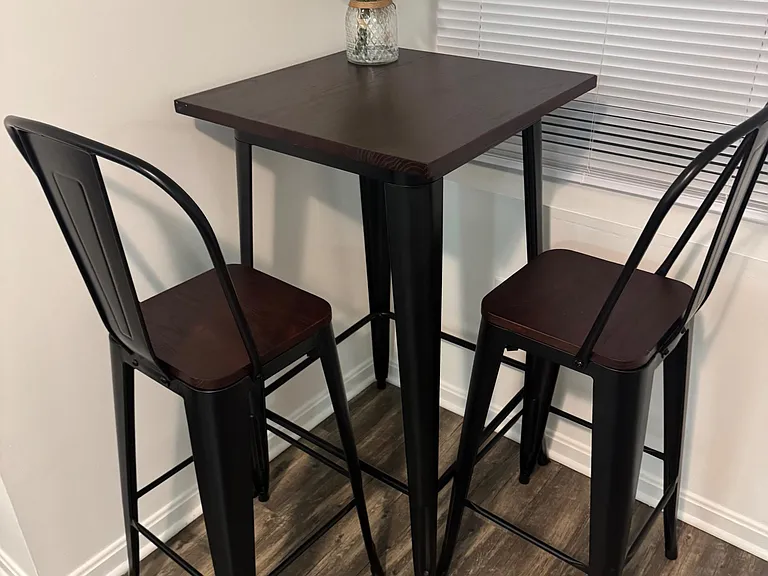 Dining nook with a tall bistro table and two bar-height chairs, set beside a window with blinds for added privacy.