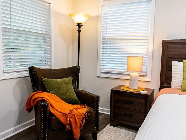 Well-furnished bedroom with a comfortable bed, nightstand lighting, and a reading chair beside dual windows with blinds.