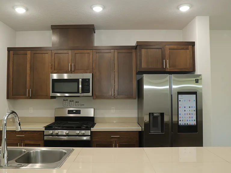 Modern kitchen with stainless steel appliances, smart touchscreen refrigerator, gas range, microwave, double_basin sink, and warm wood cabinetry. Bright recessed lighting and light countertops create a clean, contemporary look.