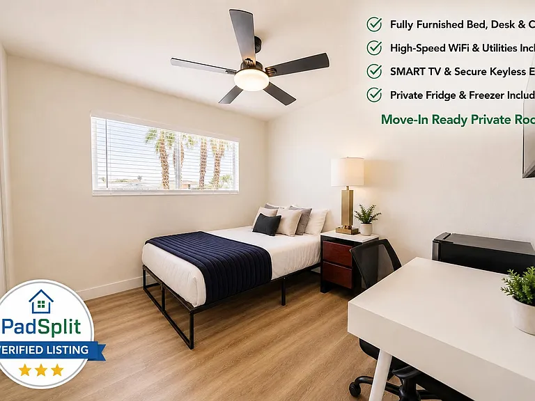 Furnished private bedroom with full bed, desk workspace, smart TV, and in-room mini fridge plus a large window.