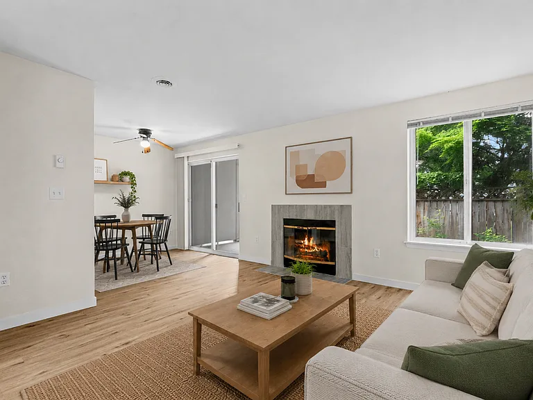 Staged living room with tile-surround fireplace, wood-style plank flooring, ceiling fan, sliding glass door patio access, and open dining area views at Garden Square Apartments in Lakewood, WA. Staged living room with tile-surround fireplace, wood-style p
