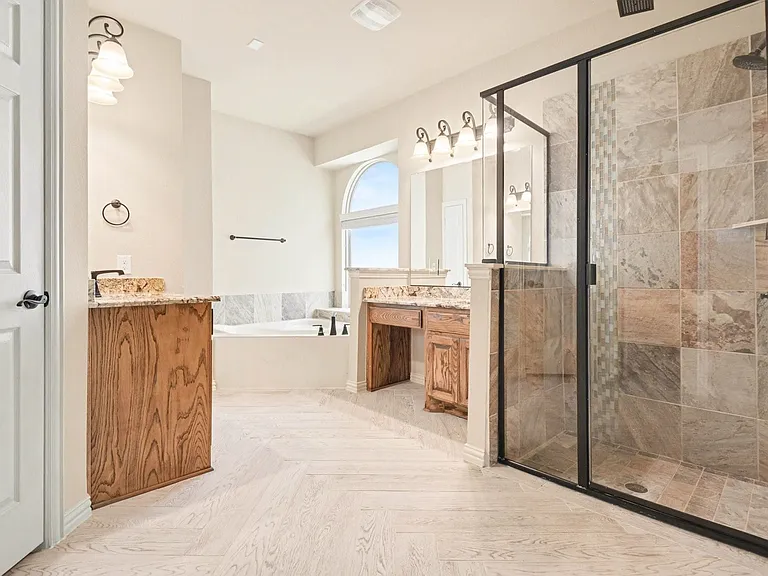 Spacious bathroom featuring light wood-finish flooring, a frameless glass shower with tile surround, a built-in soaking tub, dual vanities with stone countertops, and an arched window